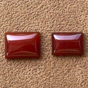 May include: Two rectangular carnelian gemstones, one larger than the other, with a deep red color and a glossy finish. The stones are set against a brown, textured background.