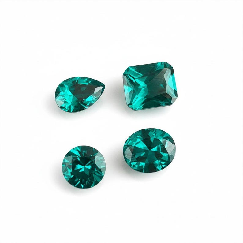 May include: Four loose faceted gemstones in a vibrant teal green color. The collection includes one pear-shaped stone, one rectangular cushion-cut stone, one round brilliant-cut stone, and one oval-cut stone, all featuring intricate geometric facets.