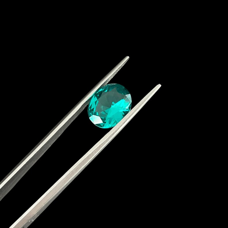 May include: A faceted oval gemstone with a vibrant teal-green color held by a pair of silver-toned metal tweezers. The gemstone displays multiple geometric facets that reflect light, creating a bright, clear appearance against a black background.