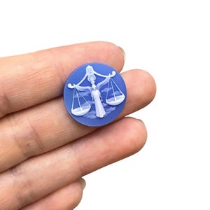 May include: A round, blue cameo featuring a white relief of a woman holding scales, representing the Libra zodiac sign. The design includes clouds and a balanced scale, set against a solid blue background. The cameo is held in a hand.