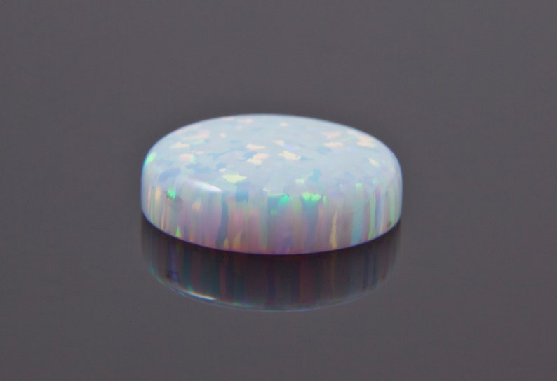 Może przedstawiać: Biały opal w szlifie kaboszonowym z iryzującymi refleksami koloru. Opal jest okrągły i ma gładką, wypolerowaną powierzchnię.