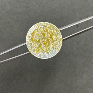 May include: A circular mother-of-pearl button featuring an iridescent yellow finish with a carved central face surrounded by leaves and a Greek key border pattern, held by a pair of silver-toned metal tweezers.