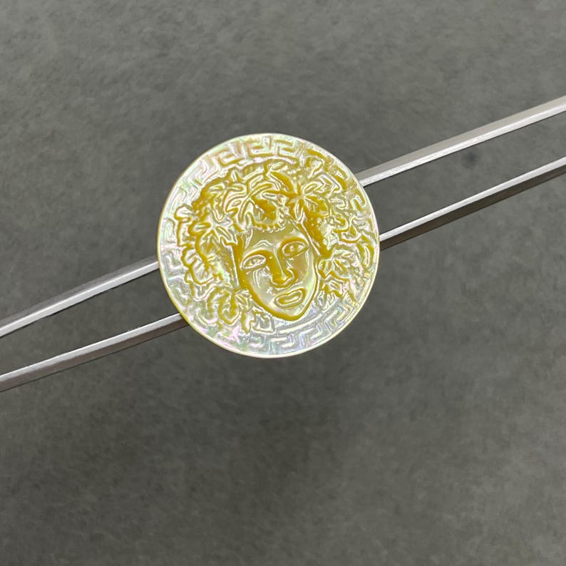May include: A circular mother-of-pearl button featuring an iridescent yellow finish with a carved central face surrounded by leaves and a Greek key border pattern, held by a pair of silver-toned metal tweezers.