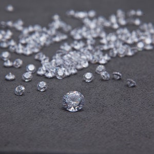May include: A close-up of a large number of clear, faceted gemstones scattered on a dark gray surface. One large gemstone is in the center of the image.