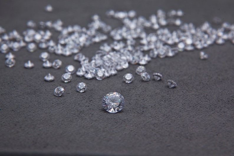 May include: A close-up of a large number of clear, faceted gemstones scattered on a dark gray surface. One large gemstone is in the center of the image.