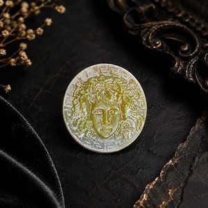 May include: A circular mother-of-pearl brooch featuring an embossed relief of a face surrounded by stylized leaves and vines. The outer edge of the disc is decorated with a geometric Greek key pattern, showcasing iridescent white and green tones.