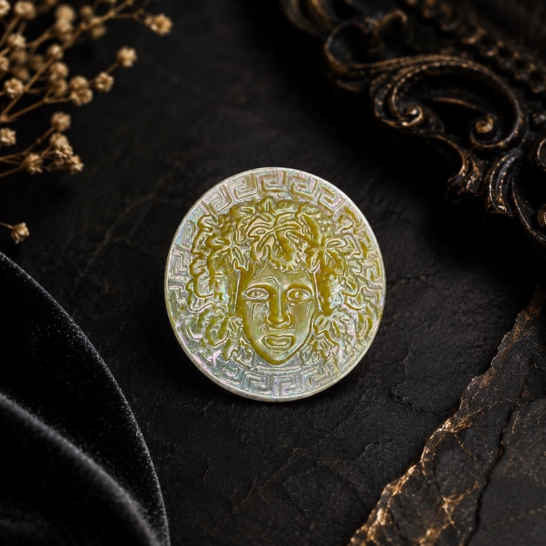 May include: A circular mother-of-pearl brooch featuring an embossed relief of a face surrounded by stylized leaves and vines. The outer edge of the disc is decorated with a geometric Greek key pattern, showcasing iridescent white and green tones.