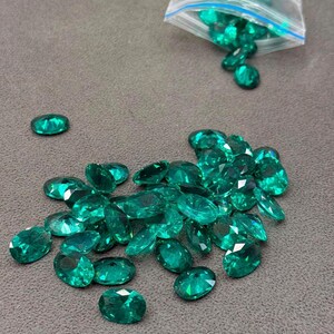 May include: A collection of loose, faceted, oval-shaped emerald green gemstones scattered across a surface. A clear plastic bag containing additional matching green gemstones is positioned at the top of the frame, with some stones spilling out.