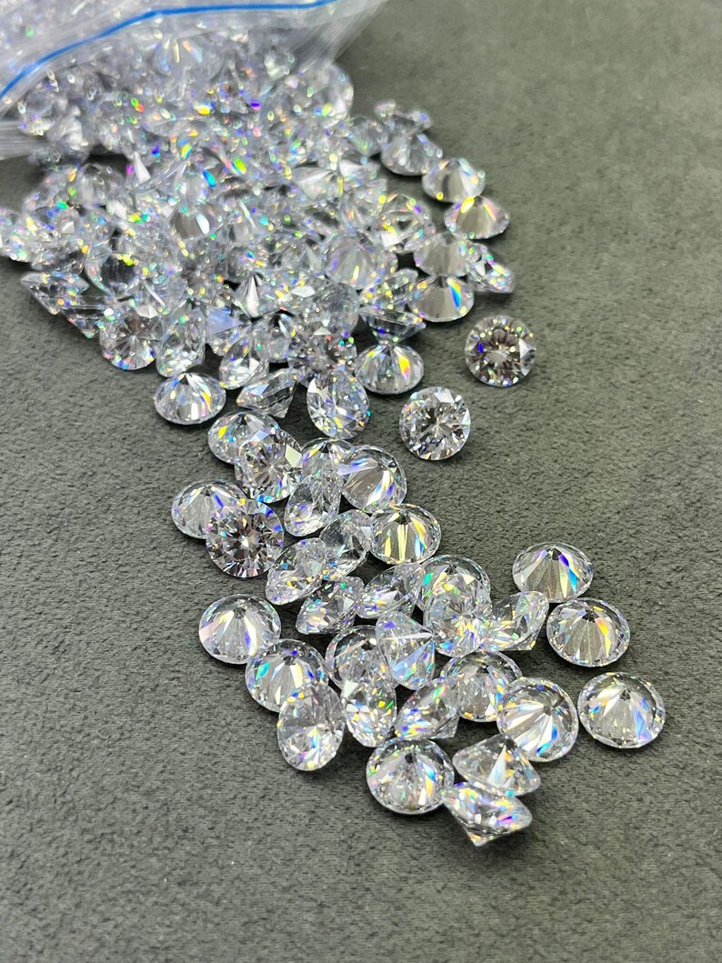 May include: A large quantity of clear, faceted, round gemstones, likely diamonds, are scattered on a gray surface. Some of the gemstones are in a clear plastic bag.