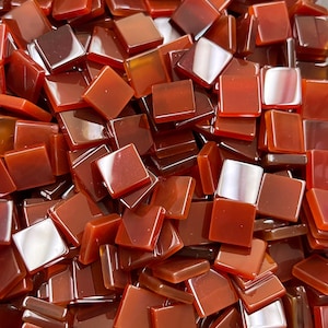 May include: A close-up view of a pile of small, square, red carnelian gemstone tiles. The tiles are smooth and shiny, with a slight variation in color and translucency.