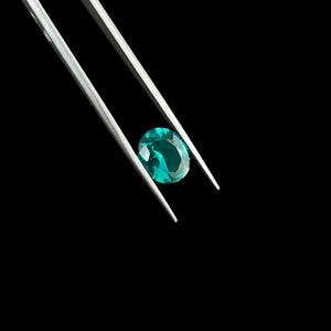 May include: A faceted oval-shaped gemstone with a vibrant teal-green color held by a pair of silver-toned metal tweezers. The gemstone displays internal light reflections and clear cut edges against a solid black background.