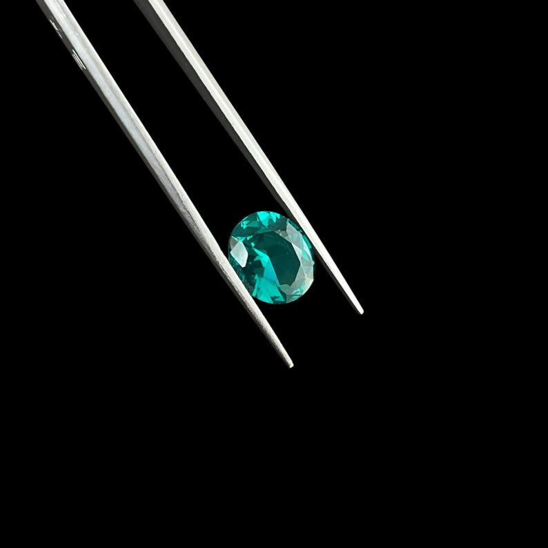 May include: A faceted oval-shaped gemstone with a vibrant teal-green color held by a pair of silver-toned metal tweezers. The gemstone displays internal light reflections and clear cut edges against a solid black background.