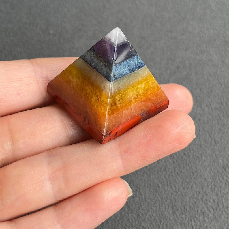 May include: A seven-layered pyramid made of various gemstones, including red, orange, yellow, white, blue, and purple. The pyramid is resting on a hand.