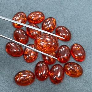 Puede incluir: Un conjunto de gemas ovaladas de color ámbar. Las piedras pulidas y brillantes se presentan sobre una superficie gris. Unas pinzas sostienen una de las piedras, revelando su transparencia y sus patrones internos, en un tono marrón-anaranjado.