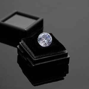 May include: A large, clear, round diamond gemstone sits in a black velvet ring box. The box is open and the diamond is centered in the box.