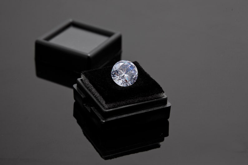 May include: A large, clear, round diamond gemstone sits in a black velvet ring box. The box is open and the diamond is centered in the box.