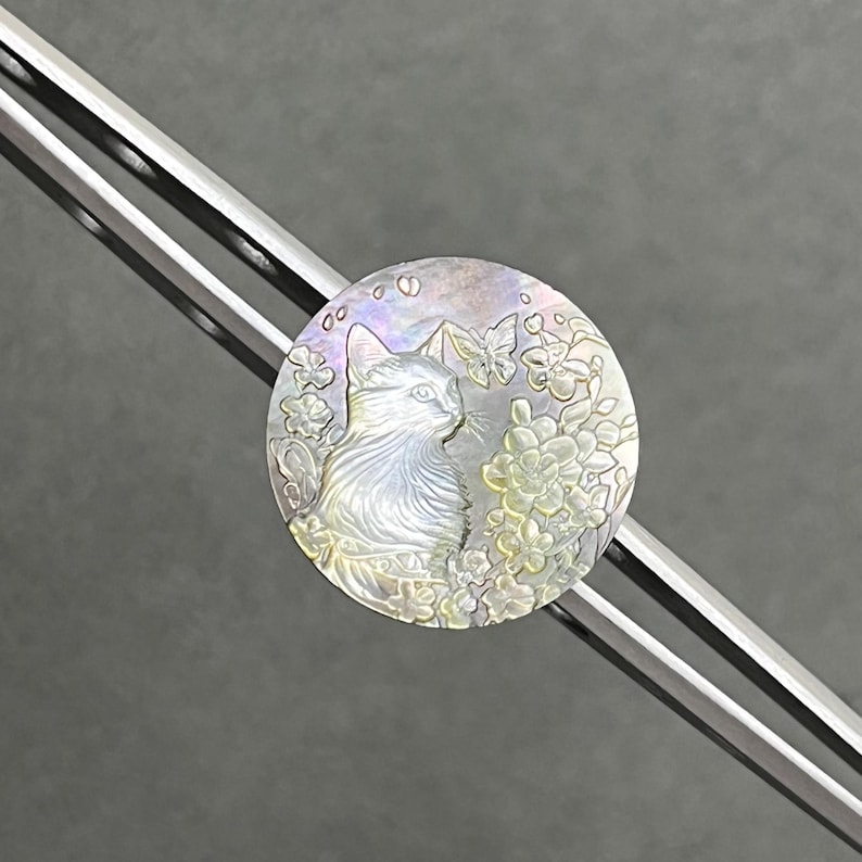 May include: A round, iridescent pendant featuring a detailed cat profile surrounded by flowers and butterflies. The pendant has a pearlescent sheen with subtle hints of color. The design is set against a gray background, with a silver bar diagonally across the image.