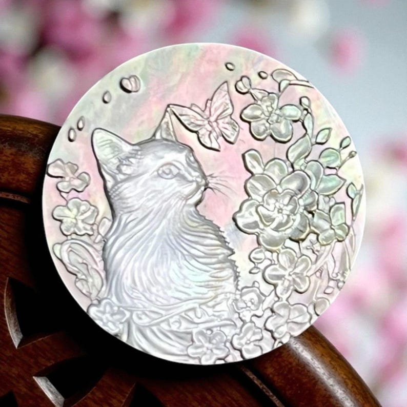 May include: A round, iridescent decorative piece featuring a cat, flowers, and a butterfly. The cat is in profile, surrounded by detailed floral and butterfly designs. The background has a soft pink and white gradient.
