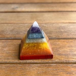 May include: A seven-layered crystal pyramid with a pointed top. The pyramid is made of clear quartz, amethyst, sodalite, clear quartz, citrine, carnelian, and red jasper. The pyramid is sitting on a wooden surface.