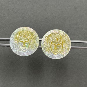 May include: A pair of circular iridescent shell buttons featuring a central relief of a face surrounded by foliage, framed by a Greek key border. The buttons are held in place by a silver-toned metal clip, showcasing a pearlescent finish.