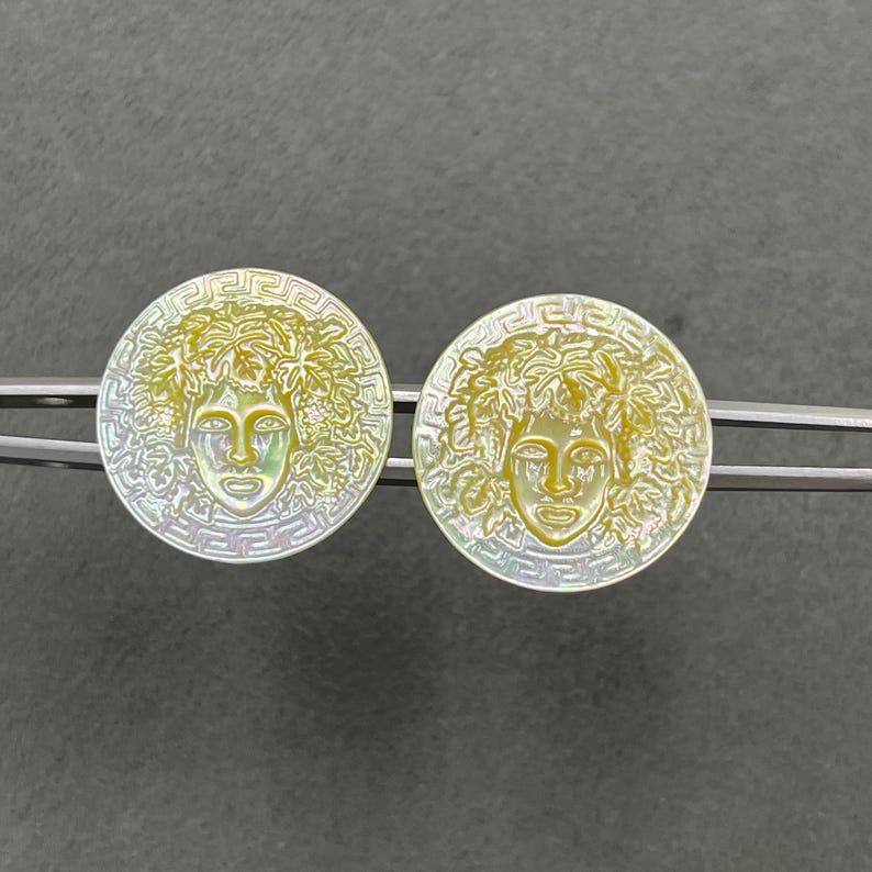 May include: A pair of circular iridescent shell buttons featuring a central relief of a face surrounded by foliage, framed by a Greek key border. The buttons are held in place by a silver-toned metal clip, showcasing a pearlescent finish.