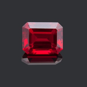 May include: A deep red gemstone with a rectangular emerald cut. The gemstone is faceted and has a high polish.