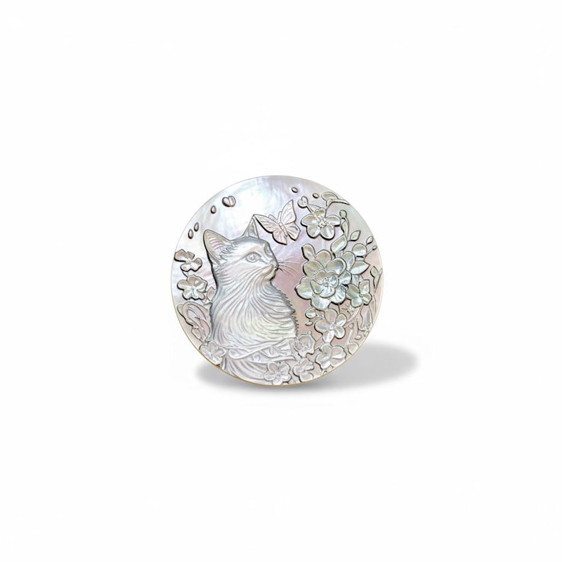 May include: A round, mother-of-pearl pendant featuring a detailed carving of a cat looking to the side. The design includes flowers and a butterfly, all in shades of white and gray. The pendant is set against a white background.