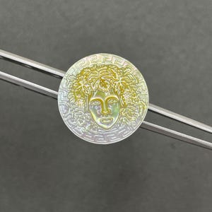 May include: A circular iridescent mother-of-pearl button featuring a central embossed face surrounded by a wreath of leaves and a Greek key border pattern, held by a pair of silver-toned metal tweezers against a dark gray background.