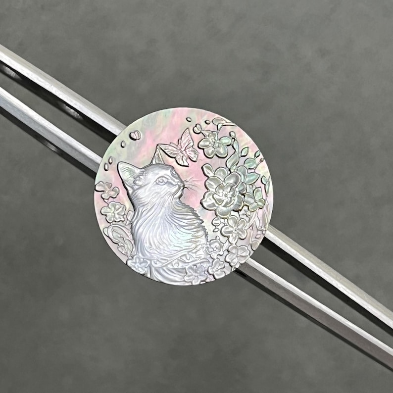 May include: A silver hair stick with a round, iridescent, mother of pearl ornament. The ornament features a carved design of a cat looking to the right, surrounded by flowers and a butterfly.