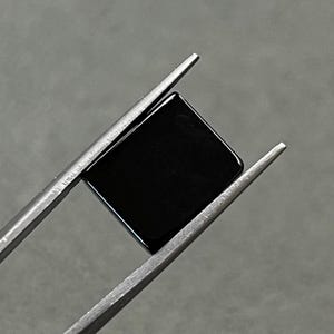 May include: A black square gemstone held by silver tweezers.