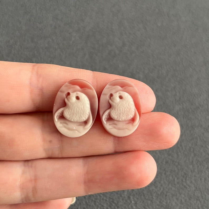 May include: Two oval-shaped cameo cabochons featuring a carved white seal figure against a soft pink background. Each piece displays a detailed relief of a seal with textured fur, rounded features, and a smooth, polished surface finish.