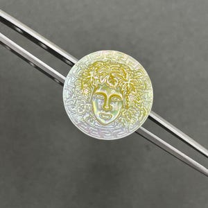 May include: A circular iridescent mother-of-pearl button featuring a central relief carving of a face surrounded by grape leaves and vines, framed by a Greek key border pattern. The button is held by a pair of silver-toned metal tweezers.