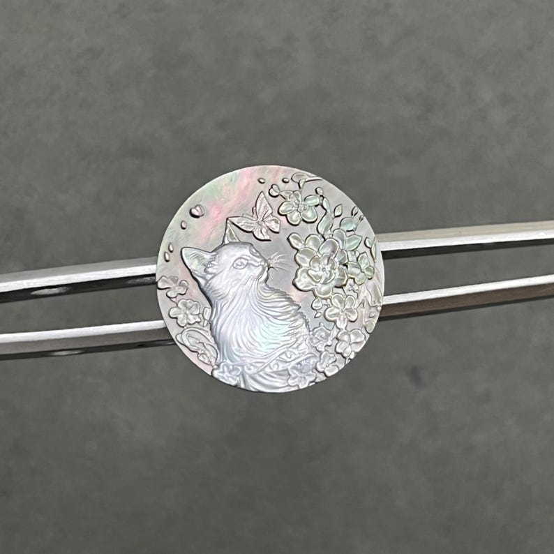 May include: A round, iridescent cat pendant with a detailed relief carving. The cat is looking up, surrounded by flowers and a butterfly. The pendant is held by a silver-colored metal bar.