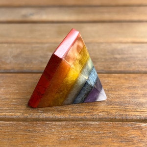 May include: A small, triangular pyramid made of layered, polished stones. The stones are arranged in a rainbow pattern, starting with red at the top and ending with purple at the bottom.