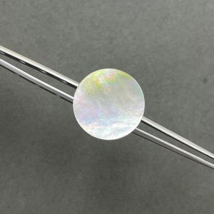 May include: A circular, iridescent mother-of-pearl inlay piece held by a pair of silver-toned metal tweezers. The inlay displays a smooth, polished surface with shifting light reflections in soft pastel tones of pink, green, and blue.