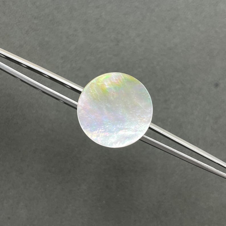 May include: A circular, iridescent mother-of-pearl inlay piece held by a pair of silver-toned metal tweezers. The inlay displays a smooth, polished surface with shifting light reflections in soft pastel tones of pink, green, and blue.