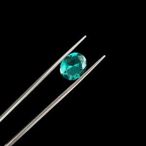 May include: A vibrant, oval-cut teal gemstone held securely between the tips of a pair of slender, silver-toned metal tweezers. The faceted stone displays a brilliant, translucent green-blue hue with sharp, geometric light reflections across its surface.