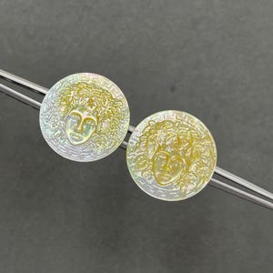May include: Two circular mother-of-pearl buttons featuring an iridescent finish and a carved relief of a face surrounded by foliage. Each button is framed by a Greek key border pattern and is held by silver-toned metal tweezers for display.