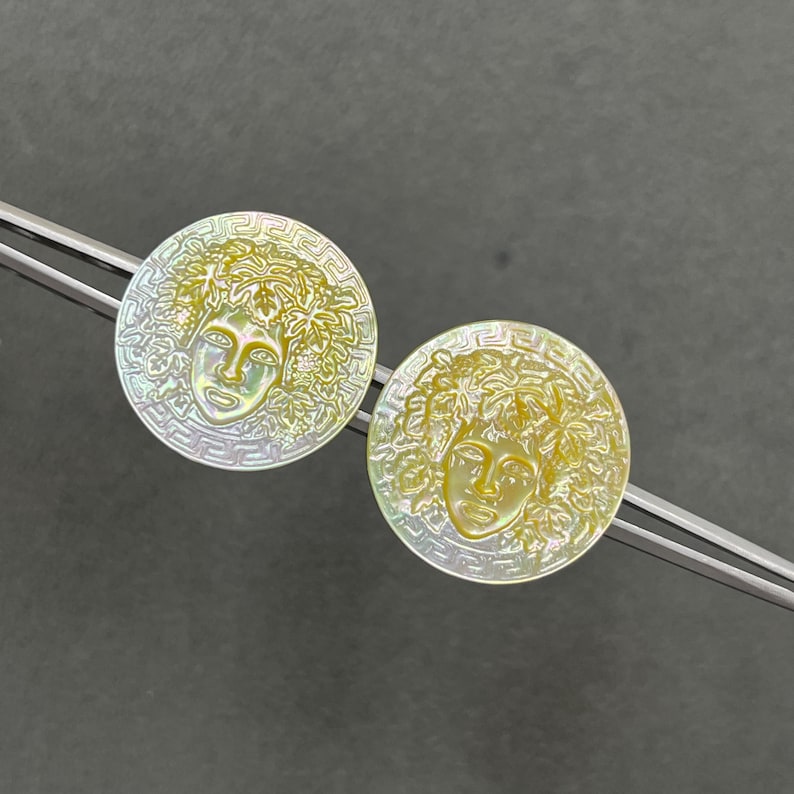 May include: Two circular mother-of-pearl buttons featuring an iridescent finish and a carved relief of a face surrounded by foliage. Each button is framed by a Greek key border pattern and is held by silver-toned metal tweezers for display.