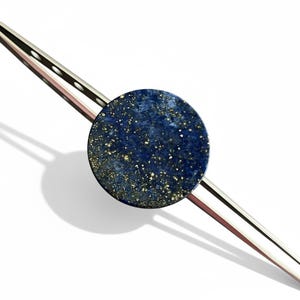 May include: A silver-toned hair clip featuring a round, dark blue stone with gold flecks. The clip has a sleek, modern design, perfect for adding a touch of elegance to any hairstyle. The stone's color and texture create a striking contrast against the silver.
