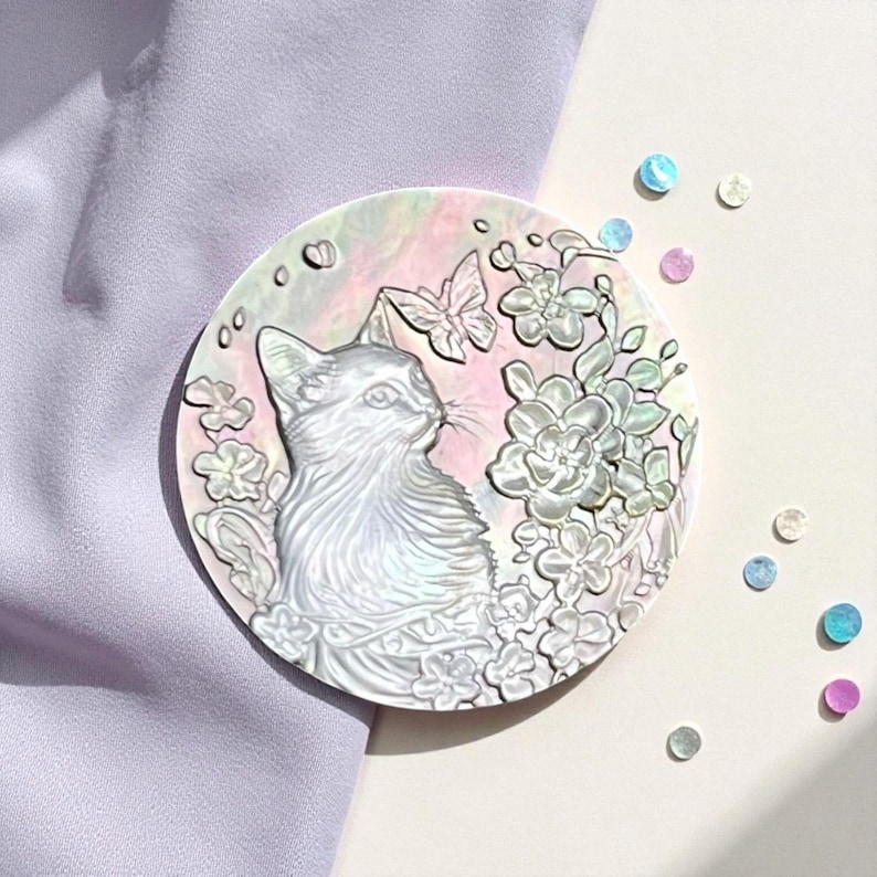 May include: A round, iridescent cat-themed decorative piece. The design features a detailed cat profile, flowers, and a butterfly, all in a pearlescent finish. The background has soft pink and white hues. Scattered iridescent sequins add a touch of sparkle.