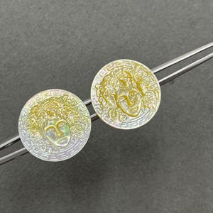 May include: Two circular iridescent mother-of-pearl buttons featuring a central embossed face surrounded by leafy hair and a Greek key border pattern. The buttons are held by silver-toned metal tweezers against a dark gray textured background.