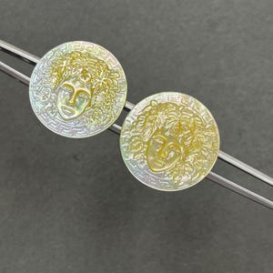 May include: Two circular mother-of-pearl buttons featuring an iridescent finish and a carved relief of a face surrounded by foliage. Each button is framed by a Greek key border pattern, showcasing a shimmering yellow and white color palette.