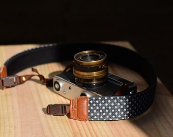 May include: A black and white patterned camera strap with brown leather accents and silver buckles. The strap is wrapped around a silver camera with a black lens.