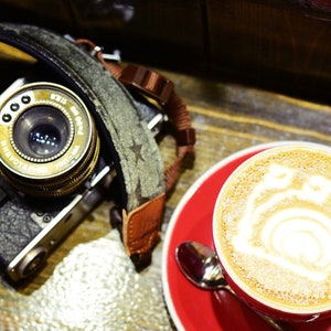 May include: A vintage camera with a leather strap sits next to a red coffee cup on a saucer. The coffee has a decorative design on top. The camera lens is gold and black, and the strap is green with star patterns.