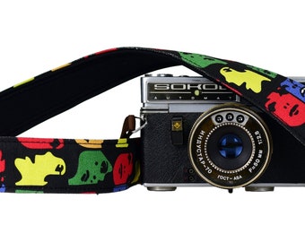 May include: A black camera strap with a colourful pattern of cartoon faces. The strap is attached to a silver and black camera with the text "Sokol Automat" on the front.
