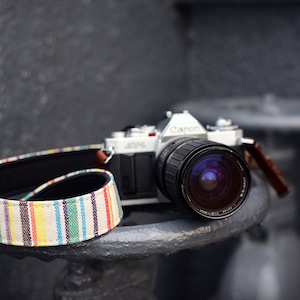 Op de afbeelding: Een zilveren Canon-camera met een zwarte lens en een kleurrijke gestreepte cameraband. De camera staat op een grijs oppervlak.