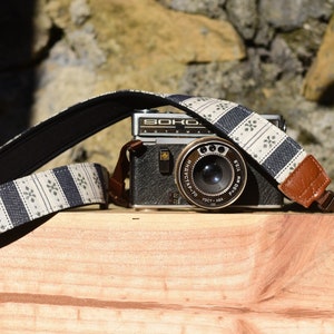 May include: A black and silver camera with a white and blue patterned camera strap. The camera has a lens with a black and silver rim and the text "SOHO" printed on the top.