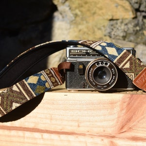 Peut inclure: Un appareil photo noir vintage avec une sangle à motifs géométriques bleus, marron et crème. L'objectif de l'appareil photo est centré et la sangle est drapée sur le corps. Le mot "SOKO" est visible sur l'appareil photo.