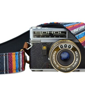 May include: A classic silver and black camera with a patterned strap. The strap has a geometric design with orange, purple, blue, and white stripes. The camera has the word "SOROL" on it.